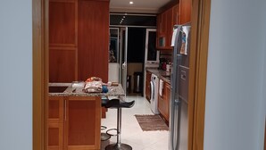 Una cocina con armarios de madera, un refrigerador de acero inoxidable, una lavadora y una zona de comedor con un taburete de bar.