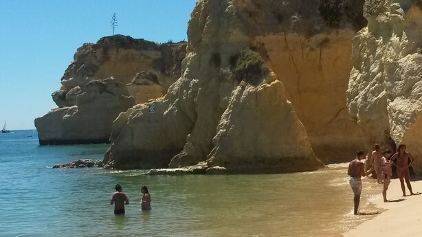 Una playa con agua cristalina, acantilados rocosos y gente nadando y caminando por la orilla.