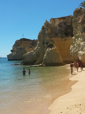 Una playa con agua cristalina, acantilados rocosos y gente nadando y caminando por la orilla.