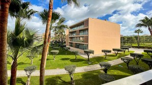 A modern building with a flat roof, surrounded by well-maintained lawns and palm trees. - Herdade dos Salgados, T2 12B_2C, Vila das Lagoas (Albufeira)