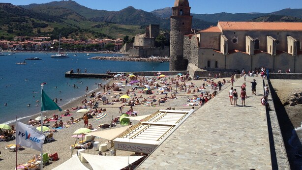 A beach with people sunbathing and swimming, a tower, and a building with a red roof.