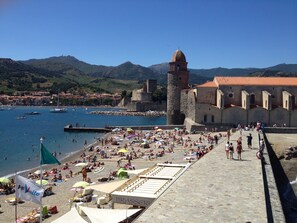 A beach with people sunbathing and swimming, a tower, and a building with a red roof. - Elegant & rustic Collioure stay. With ensuite & sea view (Collioure)