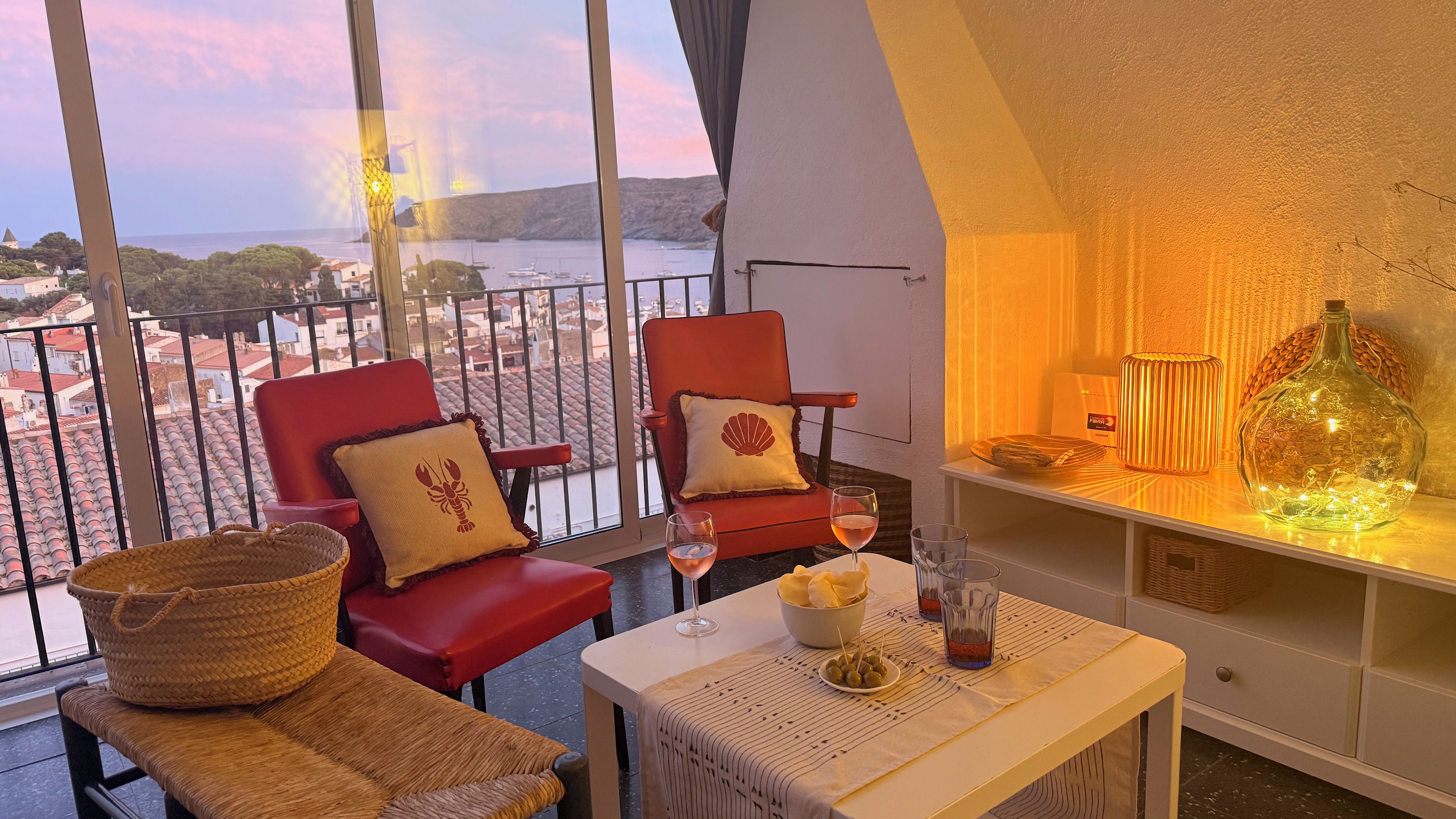 Un balcon avec vue sur la mer, des chaises rouges, une table avec un saladier rempli de collations et une bouteille de vin.