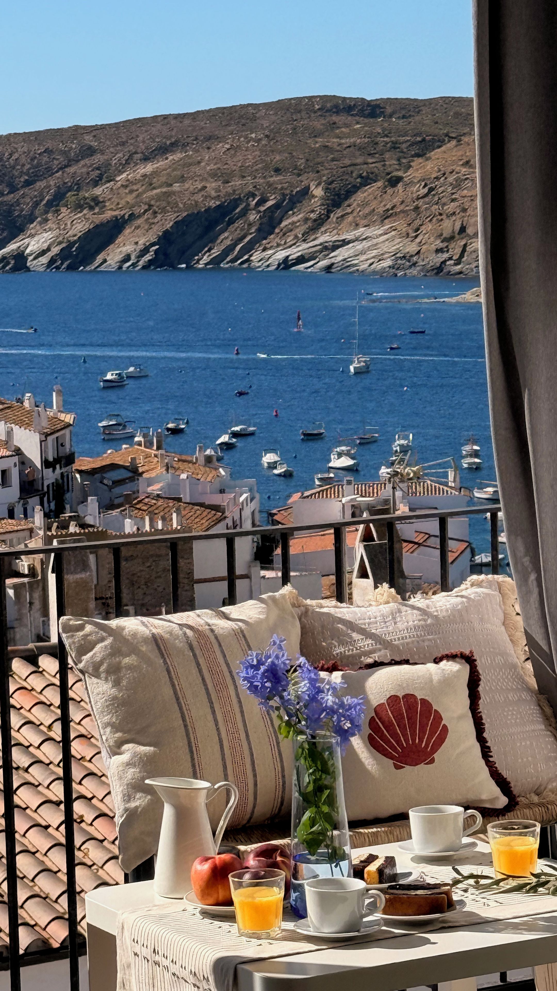 Un balcón con una mesa dispuesta para el desayuno, con vistas a un pueblo costero, barcos y colinas rocosas.