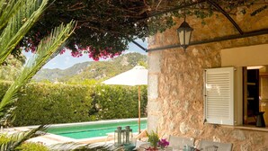 Un área de comedor junto a la piscina con una mesa para dos, pared de piedra y vista a la piscina y montañas al fondo.