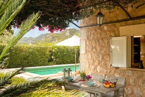 A poolside dining area with a table set for two, a stone wall, and a view of the pool and mountains in the background.