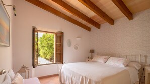 Un dormitorio con una cama grande, una ventana con contraventanas abiertas, una mesita de noche y un pequeño mueble auxiliar.