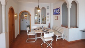 Un comedor con una mesa de madera, sillas blancas y un banco. Hay cuadros en las paredes y una lámpara colgante.