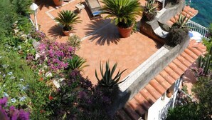 Un espacio al aire libre con terrazas, plantas en macetas y una zona de estar con vistas a un acantilado costero y al mar.