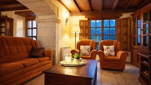 Een gezellige woonkamer met een bruine bank, twee fauteuils, een houten salontafel en een groot raam met uitzicht op bomen.