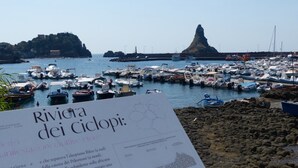 A marina with numerous boats docked, a rocky shoreline, and a sign with text in Italian. - Spacious and bright apartment with terrace and beautiful sea views (Aci Castello)