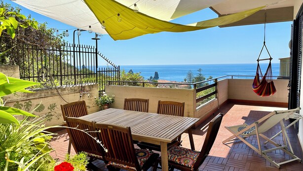 A patio with a wooden table and chairs, a hanging chair, and a view of the ocean. - Spacious and bright apartment with terrace and beautiful sea views (Aci Castello)