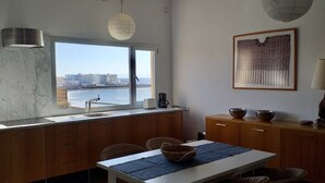 Eine Küche mit großem Fenster, das auf ein Gewässer und Gebäude blickt, Holzschränke, einem Esstisch mit Stühlen und einer Pendelleuchte.