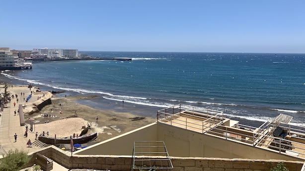 A coastal view with a beach, a pier, and a building with a balcony overlooking the sea.
