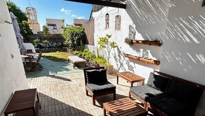A covered patio area with outdoor furniture, a small table, and a bench. There are potted plants and a hanging light fixture.