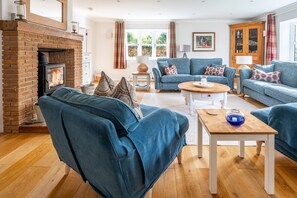 A living room with a brick fireplace, blue sofas, a wooden coffee table, and a view of greenery outside.
