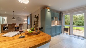 A kitchen with a wooden countertop, a dining area with wicker chairs, and a view of the garden through a glass door. - Waterside, Wiveton, Norfolk (Wiveton)