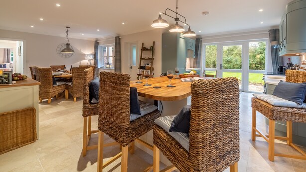 A spacious dining area with wicker chairs and a wooden table, a kitchen with blue cabinets, and large windows offering a view of the outdoors. - Waterside, Wiveton, Norfolk (Wiveton)
