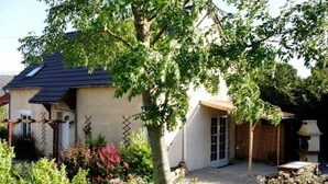 A house with a sloped roof, a garden with various plants, and a small patio area. - Family Gite in the Countryside, with Outdoor Heated Swimming Pool. Pet Friendly (Montsenelle)