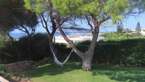 Eine Hängematze zwischen zwei Bäumen in einer grünen Umgebung mit Blick auf das Meer und Gebäude im Hintergrund.