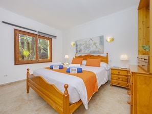 A bedroom with a wooden bed, a nightstand, a lamp, a painting, and a window with a view of trees.
