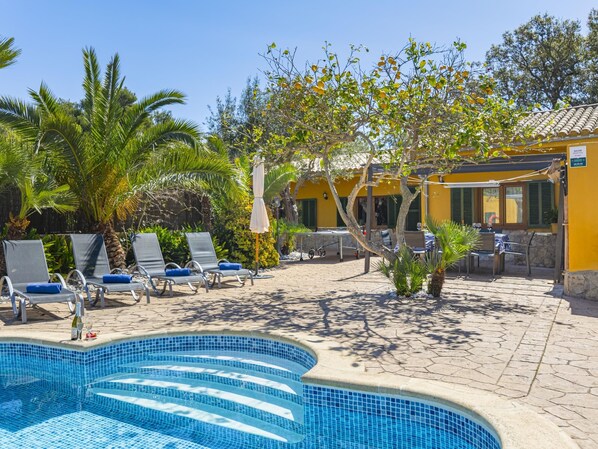 A swimming pool area with lounge chairs, a tiled deck, and a yellow building with a tiled roof.