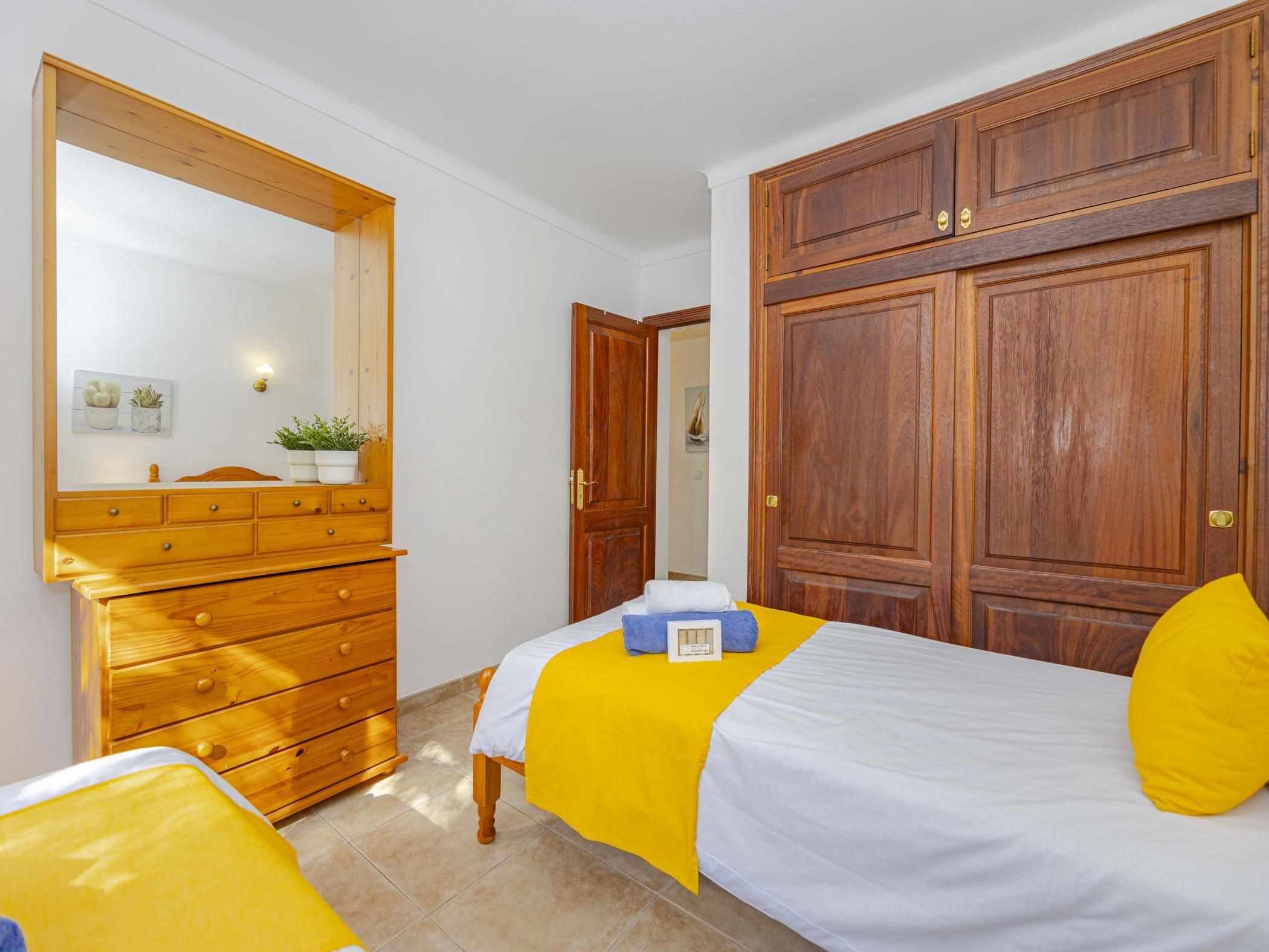 Une chambre à coucher avec une commode en bois, un lit avec une literie jaune et blanche, et une grande armoire en bois.