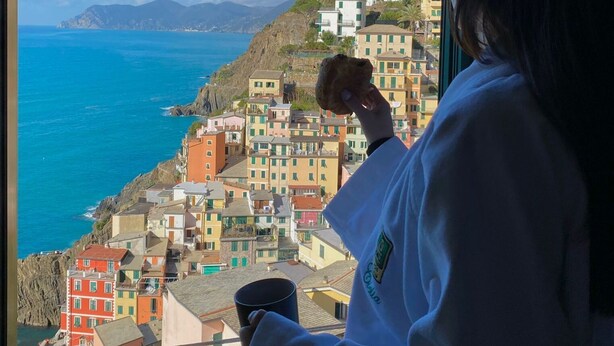 Una persona sosteniendo una taza junto a una ventana con vista a un pueblo costero de edificios coloridos y un mar azul claro.