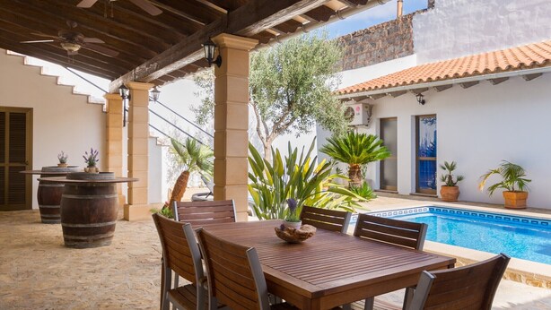 En overdækket terrasse med et træbord og stole, en pool og krukkeplanter.