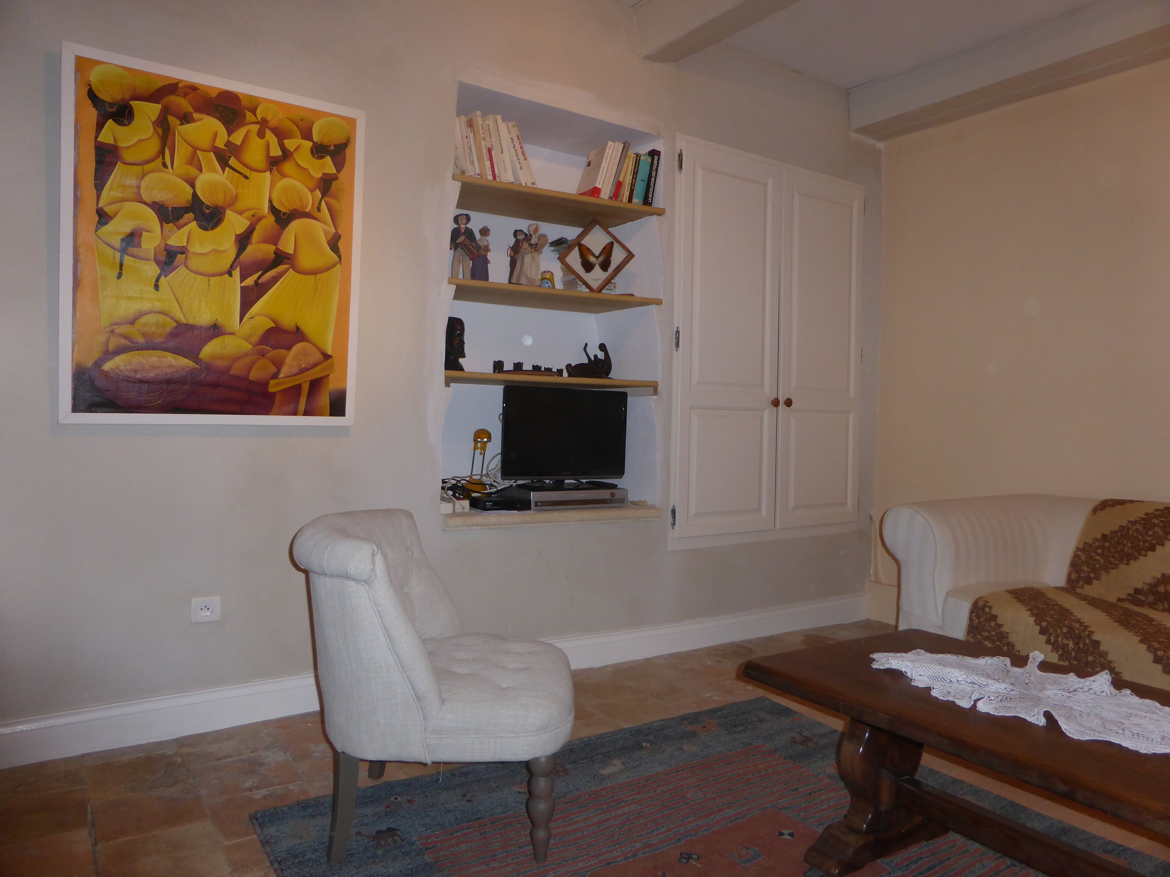 A living room with a white armchair, a wooden coffee table, a television on a shelf, and a large painting on the wall.