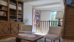 Una sala de estar con una mesa de centro de madera, dos sillones, una estantería, un ventilador y un cuadro abstracto de gran tamaño.
