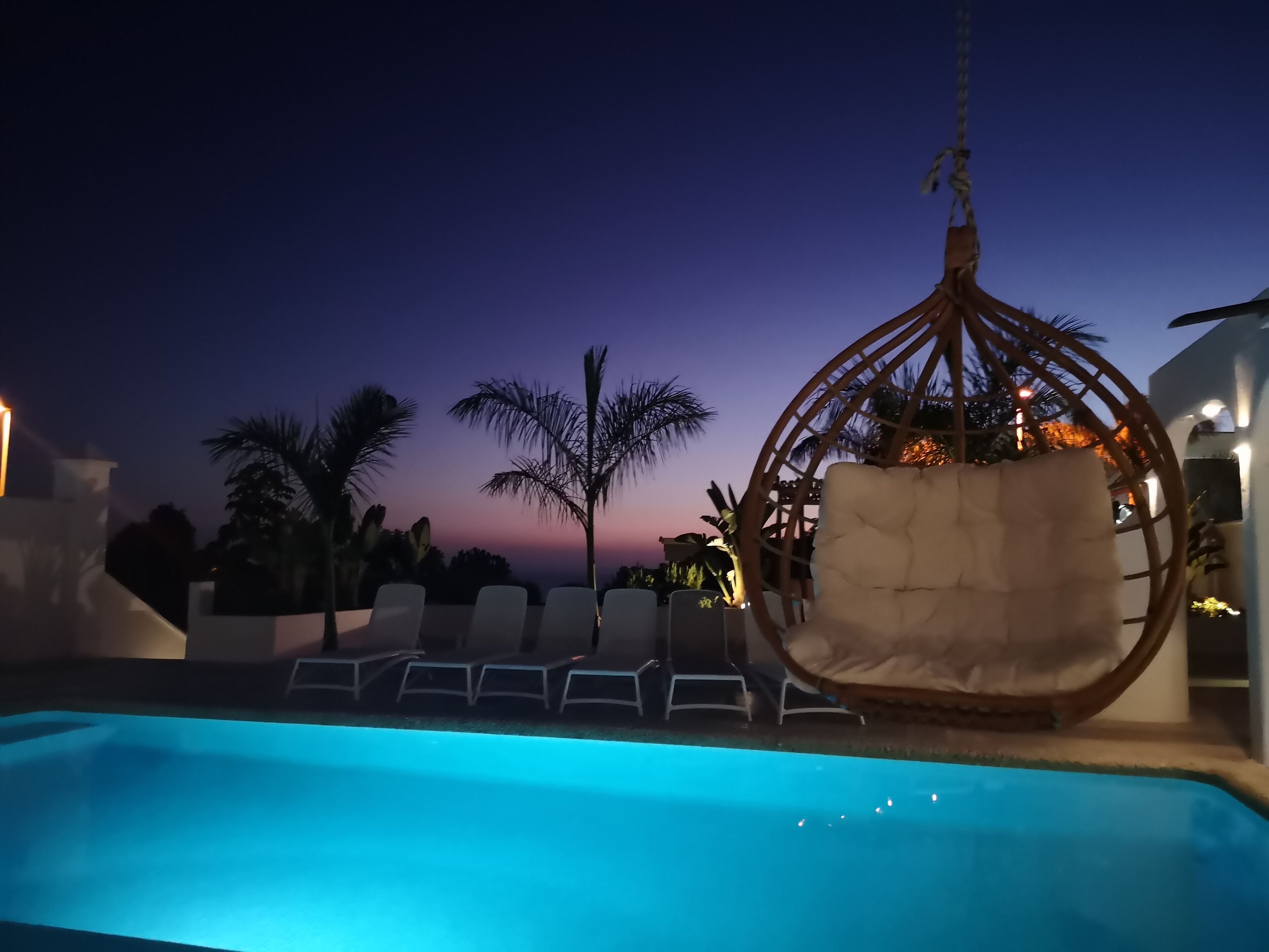 A poolside area with a hanging chair and lounge chairs, surrounded by palm trees and a sunset view.
