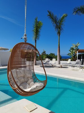 Un fauteuil suspendu au bord d’une piscine, doté de coussins blancs, entouré de chaises longues et de palmiers.