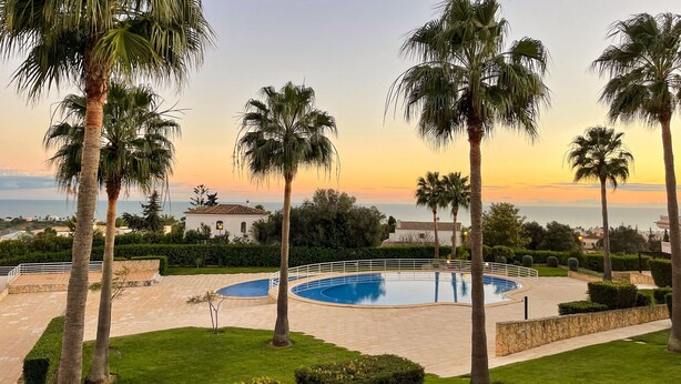 Area piscina con palme, un edificio bianco e vista sul mare al tramonto.