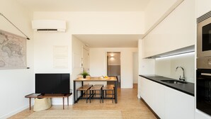 Una cocina moderna con armarios blancos, una encimera negra y un horno empotrado. Hay una zona de comedor con una mesa y sillas de madera, y un televisor a la izquierda.