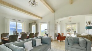 Een ruime woonkamer met een grijze bank, een eethoek met een tafel en stoelen, en een balkon met uitzicht op de bergen.