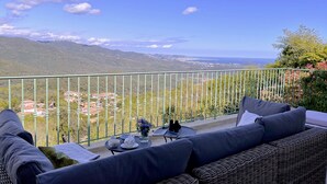 Een balkon met rieten meubels, een tafel met een vaas en uitzicht op een kustplaatsje en bergen.