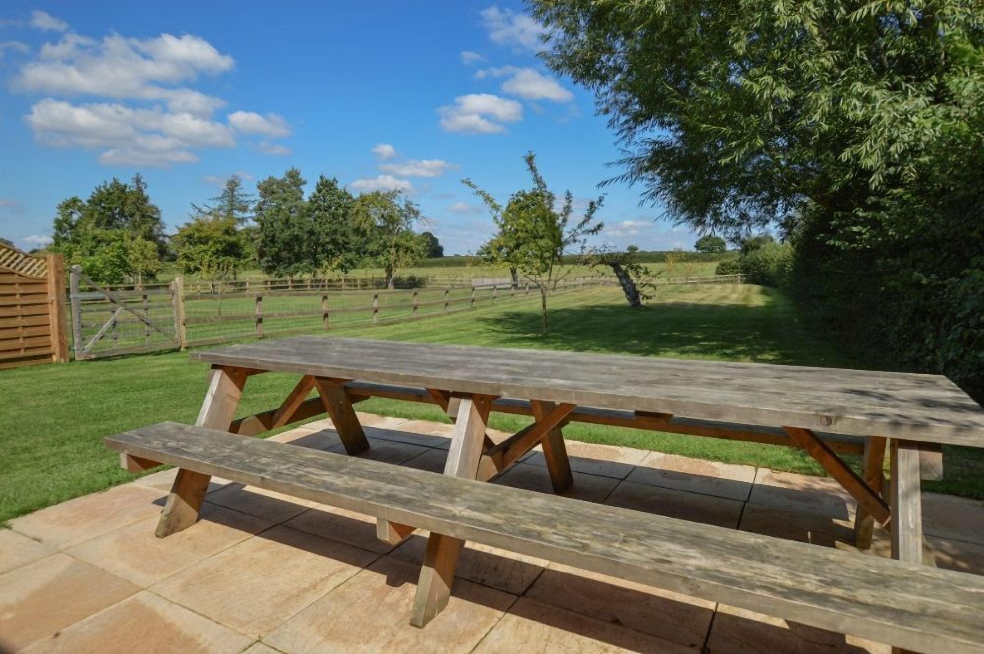 A wooden picnic table in a grassy outdoor area with a fence and trees in the background.