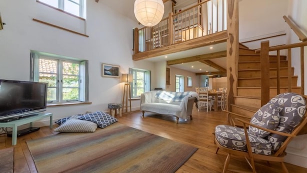 A spacious living area with a large TV, a sofa, and a patterned chair. A wooden staircase leads to an upper level with a balcony. - Field Row, Field Dalling, Norfolk (Holt)