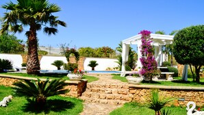 Un jardín con un sendero de piedra, palmeras y un pabellón blanco adornado con flores moradas.