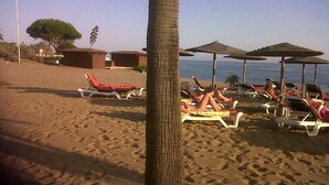 A beach with sun loungers and umbrellas, a palm tree, and a building in the background. - Stylish Luxury Apartment, Stunning Pool and Gardens, Secluded Beach (Estepona)