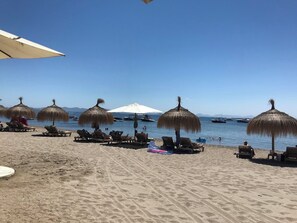 Une plage avec des transats et des parasols en chaume, un ciel dégagé et des bateaux sur l’eau.