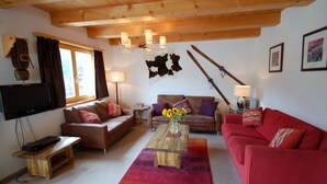 A cozy living room with a wooden ceiling, a red sofa, a brown sofa, a wooden coffee table with a vase of sunflowers, and a television on a stand.