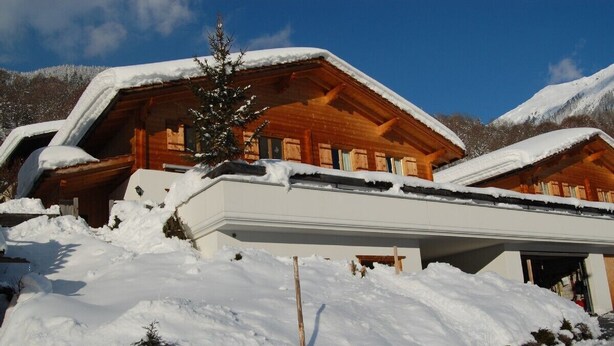 A wooden chalet with a sloped roof covered in snow, surrounded by a snowy landscape and mountains.
