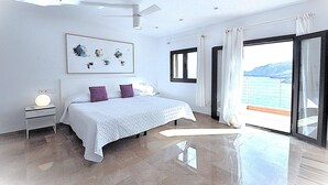 Une chambre moderne avec un grand lit, un ventilateur de plafond, une table de chevet avec une lampe et une vue sur la mer, visible à travers une porte-fenêtre coulissante.