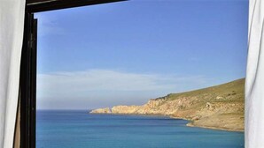 Une vue sur la côte, aperçue à travers une fenêtre ornée de rideaux, avec un ciel dégagé, une mer calme et des falaises rocheuses.