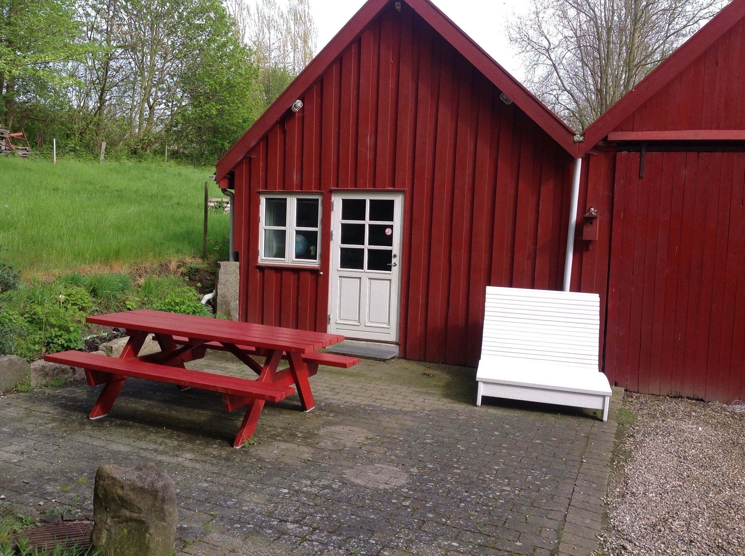 A red wooden building with a white door and windows, a picnic table, and a white bench outside.