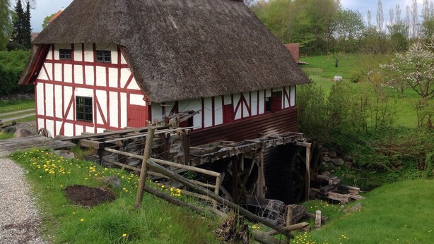 A traditional half-timbered house with a thatched roof, situated next to a watermill.