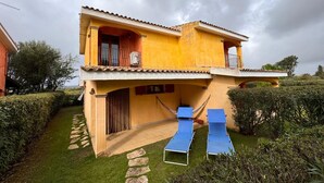 A two-story yellow building with a balcony, a small garden, and two blue lounge chairs.