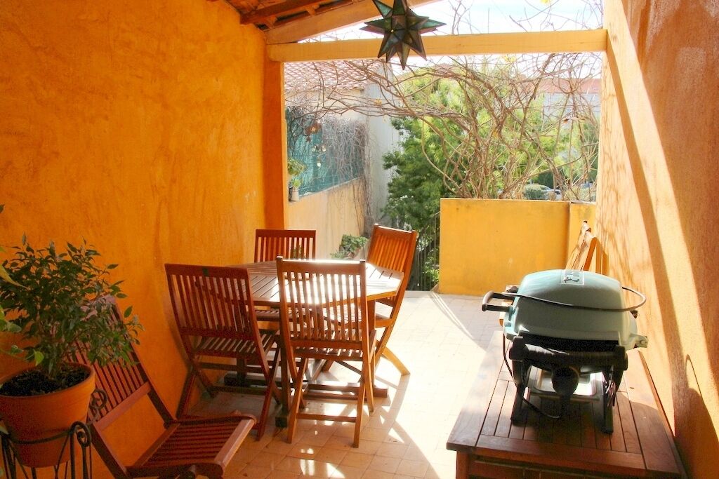 An outdoor patio with a wooden table and chairs, a grill, and a potted plant.